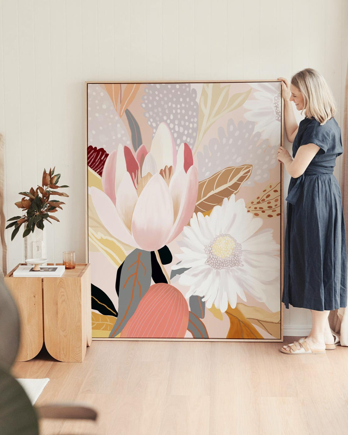 A woman standing next to a framed canvas print depicting a floral scene with soft, warm tones.