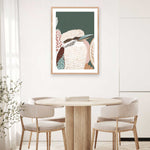 A framed print of a kookaburra perched on a branch, with lush green tones in the background, displayed on a wall above a circular wooden table and flanked by two chairs with a plant to the side.