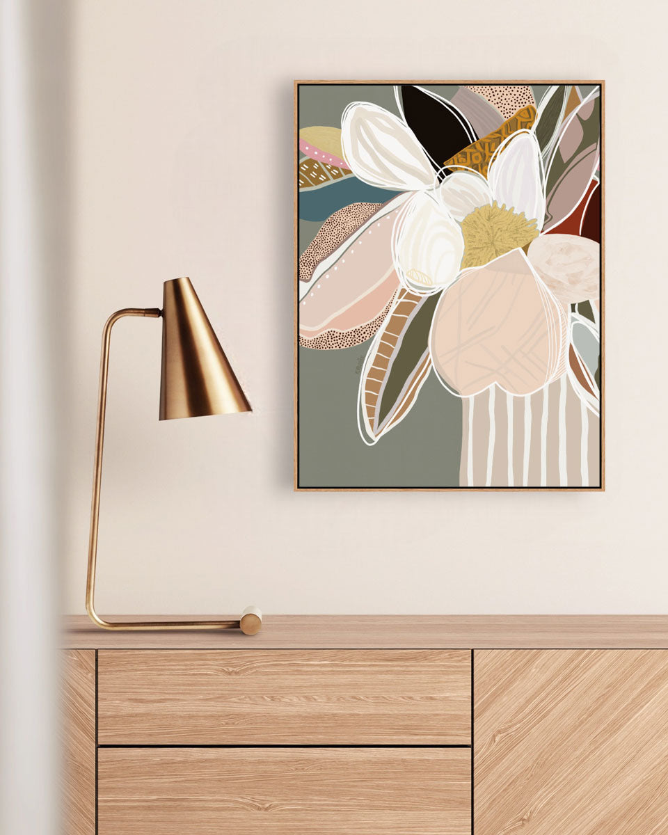 A framed canvas print titled 'Elodie In Sage No.2' hanging on a wall above a wooden cabinet, accompanied by a gold lamp with a white shade to the left.