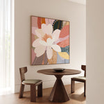 A framed canvas print titled 'Flourish', depicting a vibrant floral scene with a large flower in the center, displayed on a wall above a circular wooden table and flanked by two chairs.
