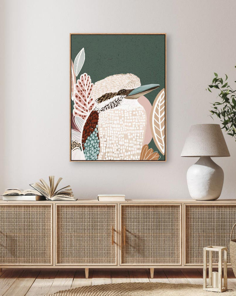 A framed canvas print of a kookaburra bird with a lush green background, displayed in a room setting with a wooden cabinet and a white lamp.