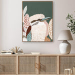 A framed canvas print of a kookaburra bird with a lush green background, displayed in a room setting with a wooden cabinet and a white lamp.