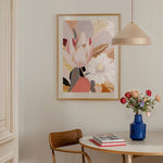 A framed abstract floral print hanging on a wall above a round table, with a chair and a pendant lamp in the room.