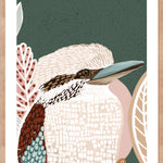A framed print of a kookaburra perched on a branch, with lush green tones in the background, displayed on a wall above a circular wooden table and flanked by two chairs with a plant to the side.