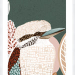 A framed print of a kookaburra perched on a branch, with lush green tones in the background, displayed on a wall above a circular wooden table and flanked by two chairs with a plant to the side.