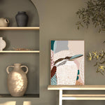A framed canvas print titled 'Kookaburra in Pink No.2' displayed on a white wall, featuring an abstract depiction of a kookaburra with soft pink and beige tones.