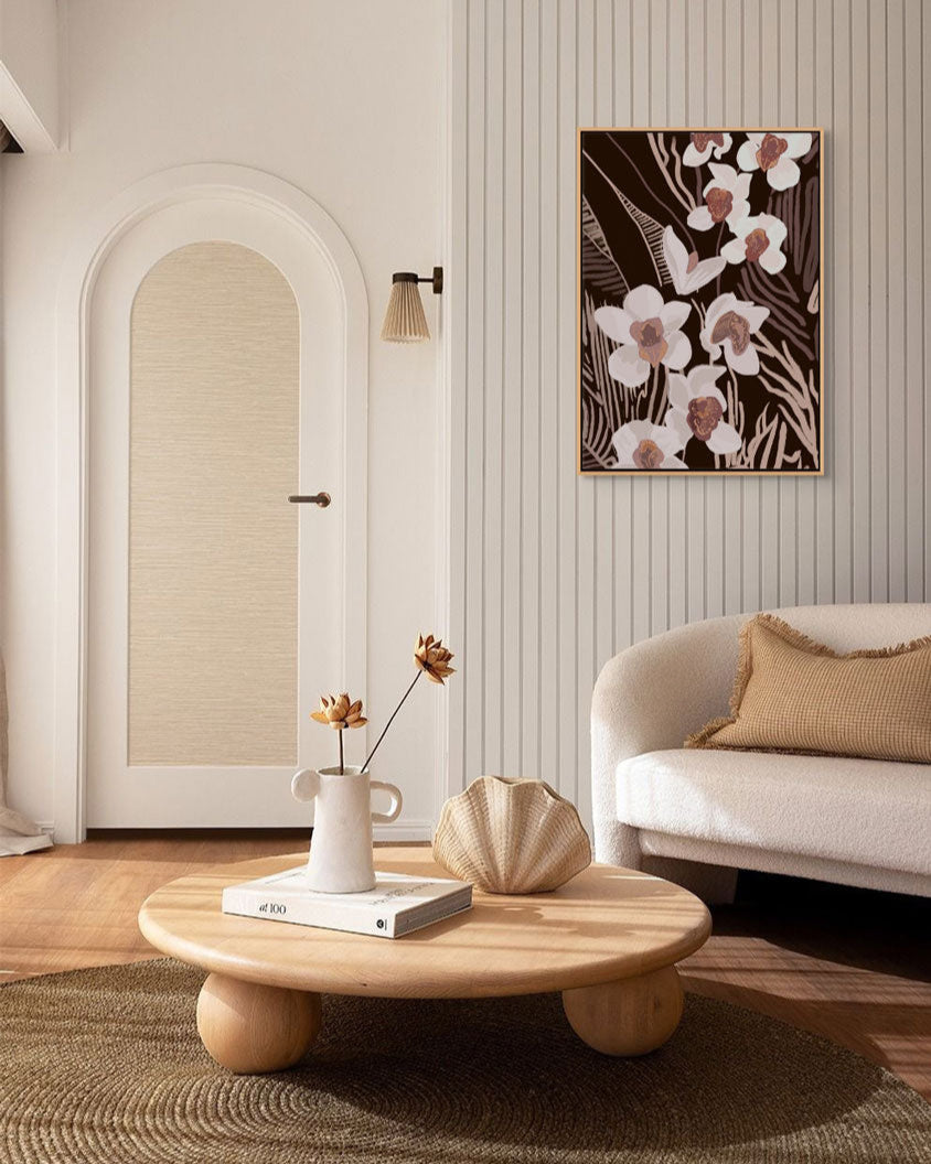 A framed canvas print titled 'The Light Beneath No.2', depicting a floral pattern with soft illumination, hung on a wall above a white sofa, with a round wooden table in the foreground containing a vase with flowers and a decorative shell.
