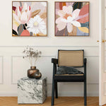 Two framed canvas prints featuring floral and abstract designs, hanging on a wall above a small marble-topped side table and next to a black chair with a cushion on it.