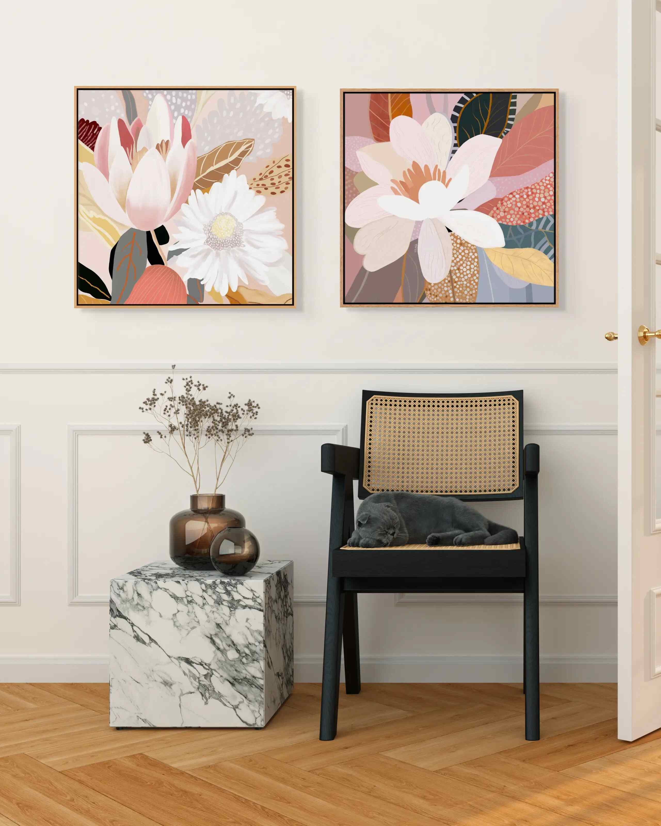 Two framed canvas prints featuring floral and abstract designs, hanging on a wall above a small marble-topped side table and next to a black chair with a cushion on it.