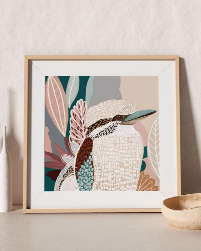 A framed print of a kookaburra surrounded by abstract pink shapes and leaves, displayed on a table with a light wooden frame.