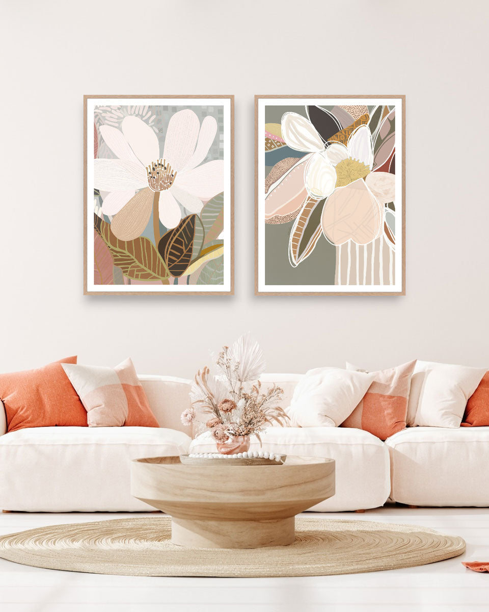 Two framed art prints featuring abstract floral designs hanging on a wall over a white sofa, with orange cushions, a wooden table, and a decorative bowl in the foreground.