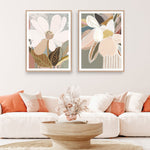Two framed art prints featuring abstract floral designs hanging on a wall over a white sofa, with orange cushions, a wooden table, and a decorative bowl in the foreground.