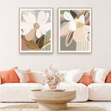 Two framed art prints featuring abstract floral designs hanging on a wall over a white sofa, with orange cushions, a wooden table, and a decorative bowl in the foreground.