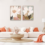 Two framed canvas prints featuring floral artwork, hanging on a wall above a white sofa, with one print slightly to the left and the other to the right. The room is styled with neutral tones and has a modern aesthetic.