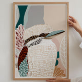 Framed artwork of a kookaburra with abstract patterns held by a person against a white wall.