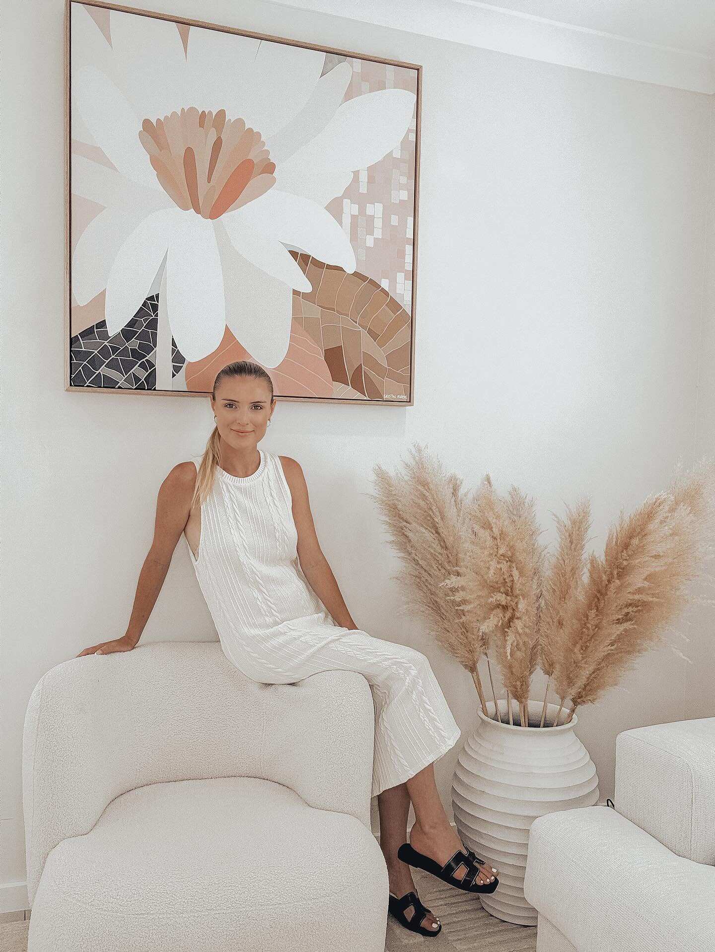 A woman sitting on a white surface in a minimalist white room with a large framed canvas print of a floral piece in the background and a potted plant with tall beige grasses to the side.
