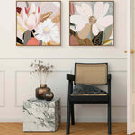 Two framed canvas prints featuring floral artwork, hanging on a wall above a small marble-topped side table and a black chair.