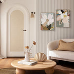 A framed canvas art piece with floral print hanging on a wall above a white sofa, with a round wooden coffee table in the foreground featuring a book and a vase with flowers.