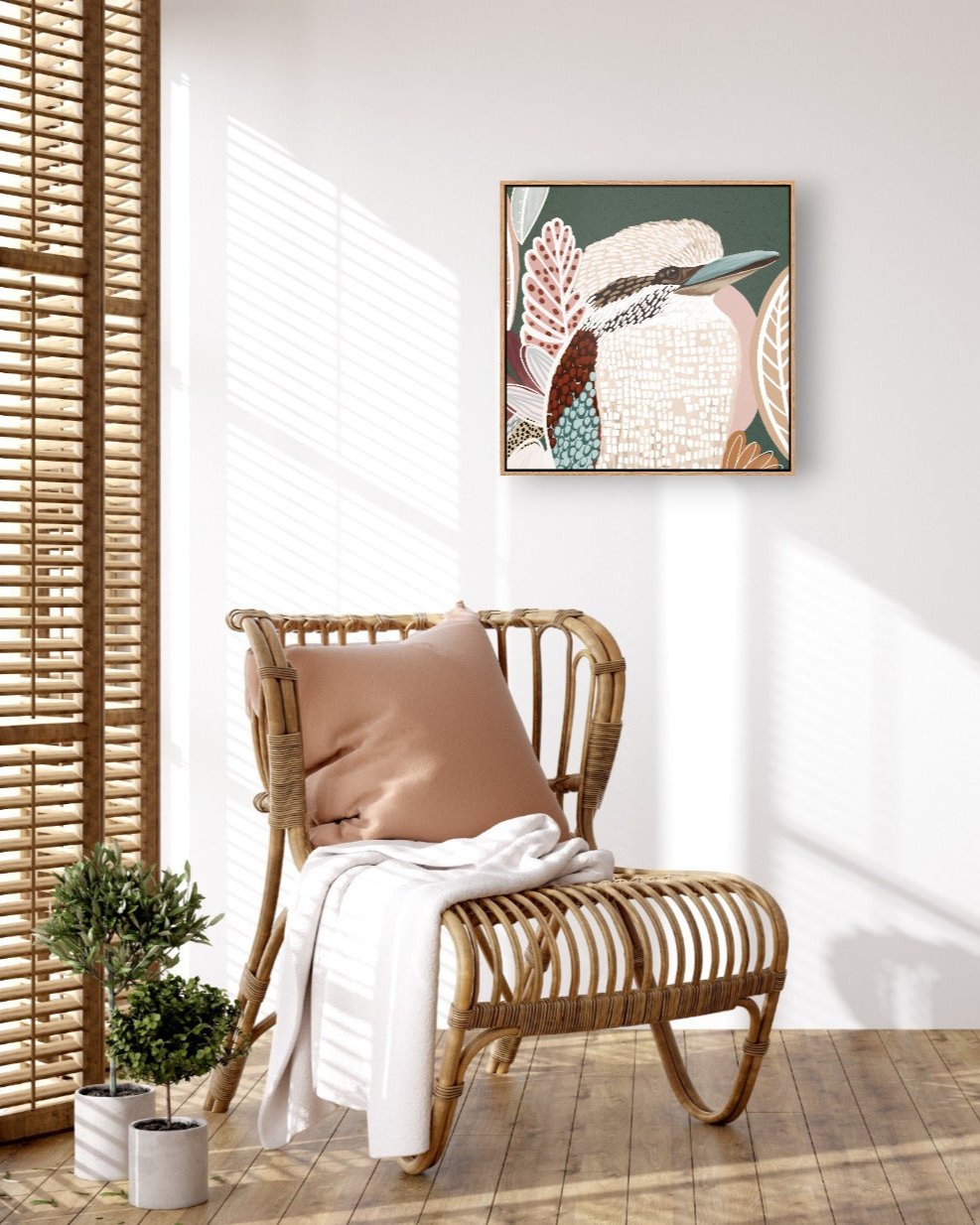 A framed canvas print titled 'Kookaburra in Green' displayed on a wall above a wooden chair with a pillow and a white cloth, with a small potted plant to the side.
