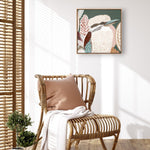 A framed canvas print titled 'Kookaburra in Green' displayed on a wall above a wooden chair with a pillow and a white cloth, with a small potted plant to the side.