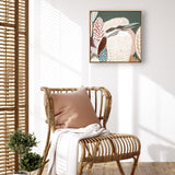 A framed canvas print titled 'Kookaburra in Green' displayed on a wall above a wooden chair with a pillow and a white cloth, with a small potted plant to the side.