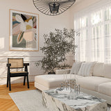 A framed print titled 'Elodie Sage' displayed on a wall above a neutral-colored sofa in a well-lit room with a large window, a decorative hanging light, and a coffee table with a plant.