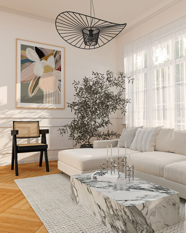 A framed print titled 'Elodie Sage' displayed on a wall above a neutral-colored sofa in a well-lit room with a large window, a decorative hanging light, and a coffee table with a plant.