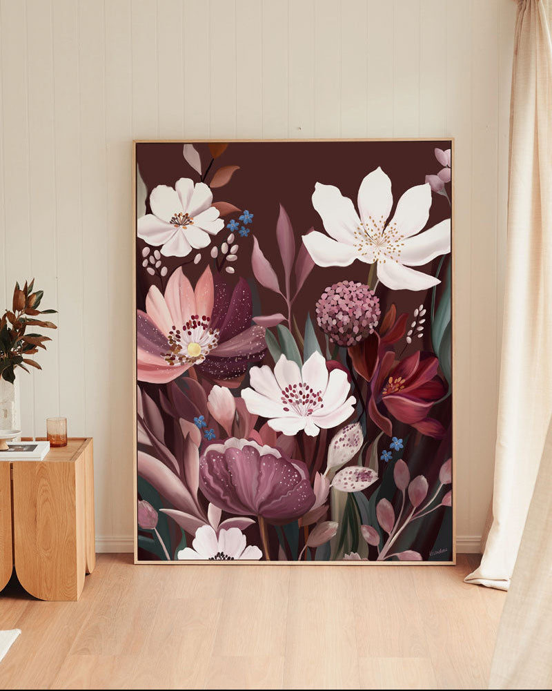A large framed botanical art print featuring white and pink flowers with deep purple and burgundy tones, leaning against a beige wall in a modern room with light wood flooring.