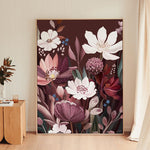A large framed botanical art print featuring white and pink flowers with deep purple and burgundy tones, leaning against a beige wall in a modern room with light wood flooring.