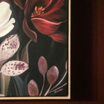 A framed floral painting hanging on a dark wood wall, featuring red, pink, white, and purple flowers against a dark background.