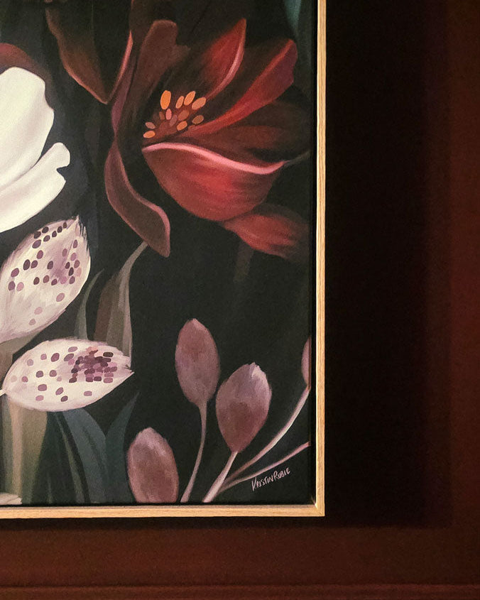 A framed floral painting hanging on a dark wood wall, featuring red, pink, white, and purple flowers against a dark background.