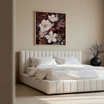 A framed floral wall art print hangs above a modern platform bed with white bedding in a softly lit beige bedroom. The artwork features large cream, white, and dusty pink flowers with dark plum and brown botanical details, set in a thin light wood frame.