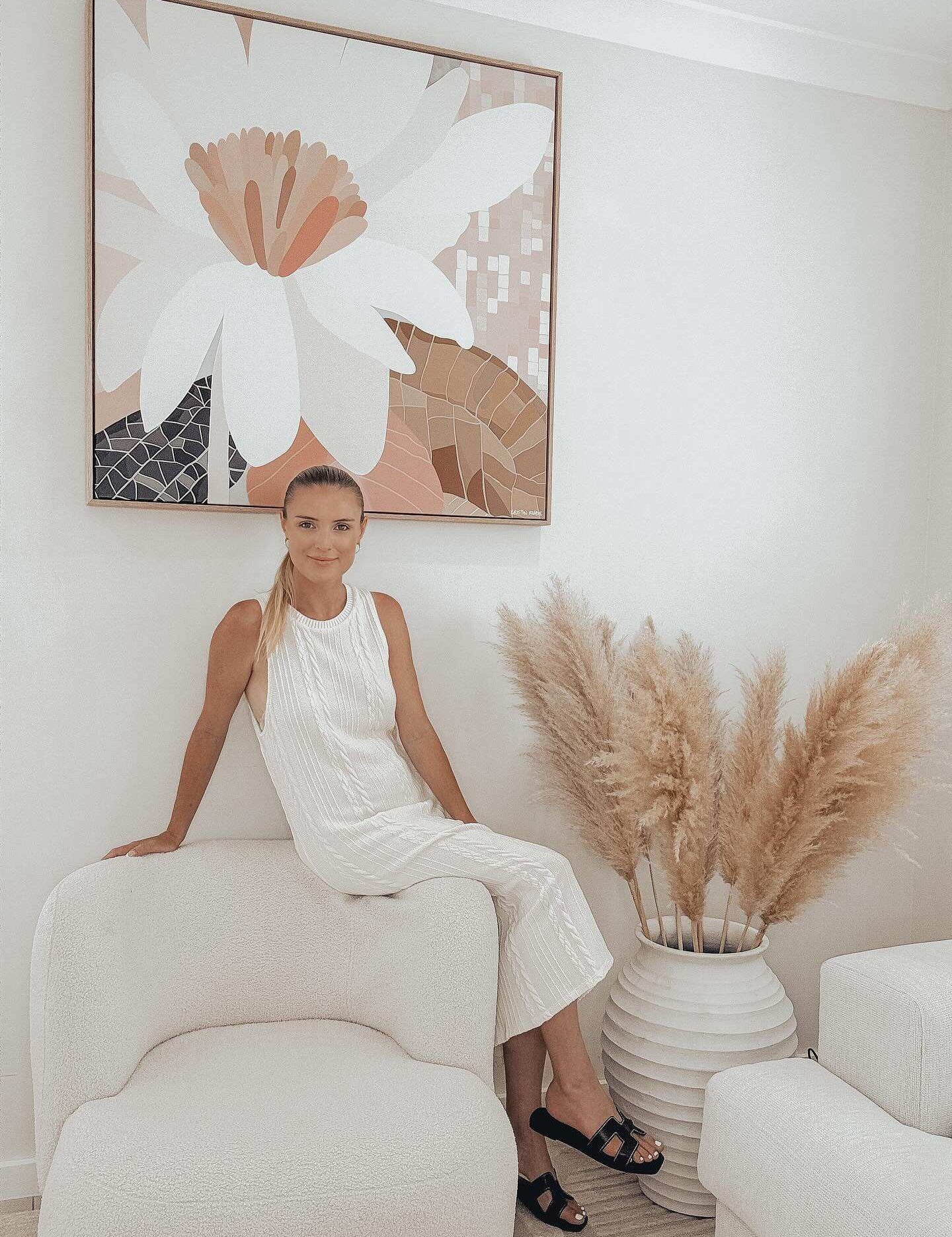 A woman sitting on a white surface in a minimalist white room with a large framed canvas print of a floral piece in the background and a potted plant with tall beige grasses to the side.