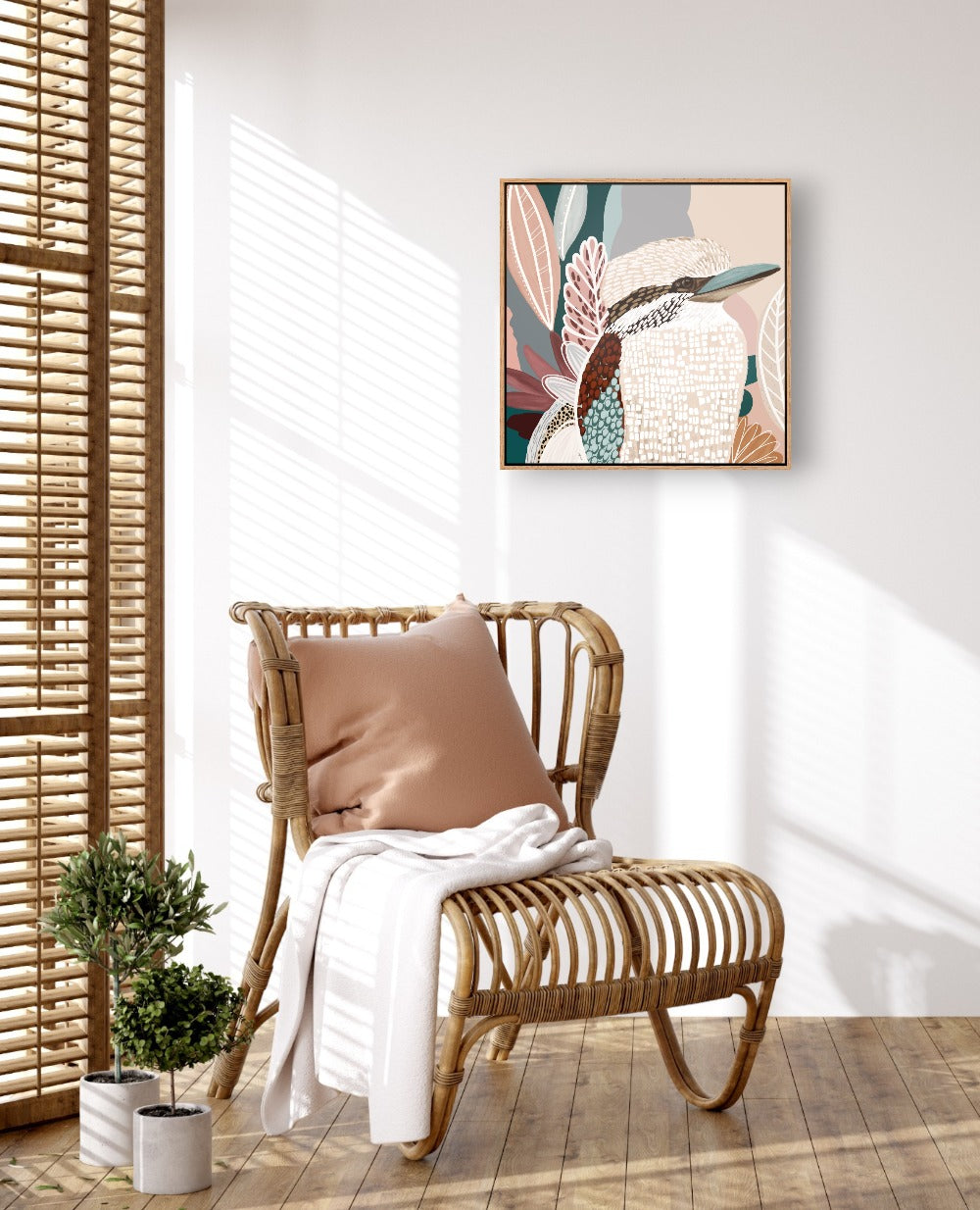 Framed canvas print of a Kookaburra perched on a branch with pink blossoms in the background, displayed above a rattan chair with a white cushion and a small potted plant.